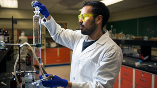 A male student in a chemistry lab is pouring fluid from one beaker into a larger test tube.